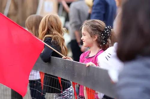 Un enfant tient un drapeau rouge et observe une activité depuis derrière une clôture. 