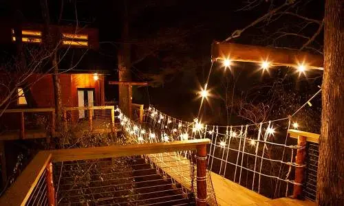 Un pont en bois vers une cabane dans les arbres éclairée par des guirlandes lumineuses