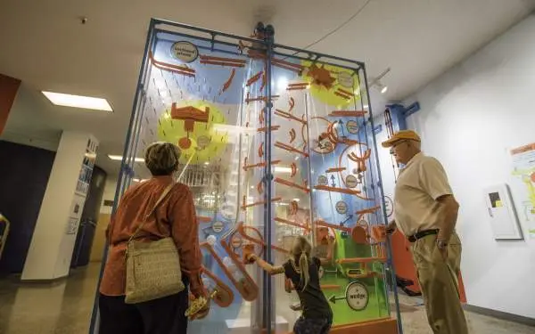 Un enfant joue avec une exposition interactive dans un musée, sous le regard de ses grands-parents.