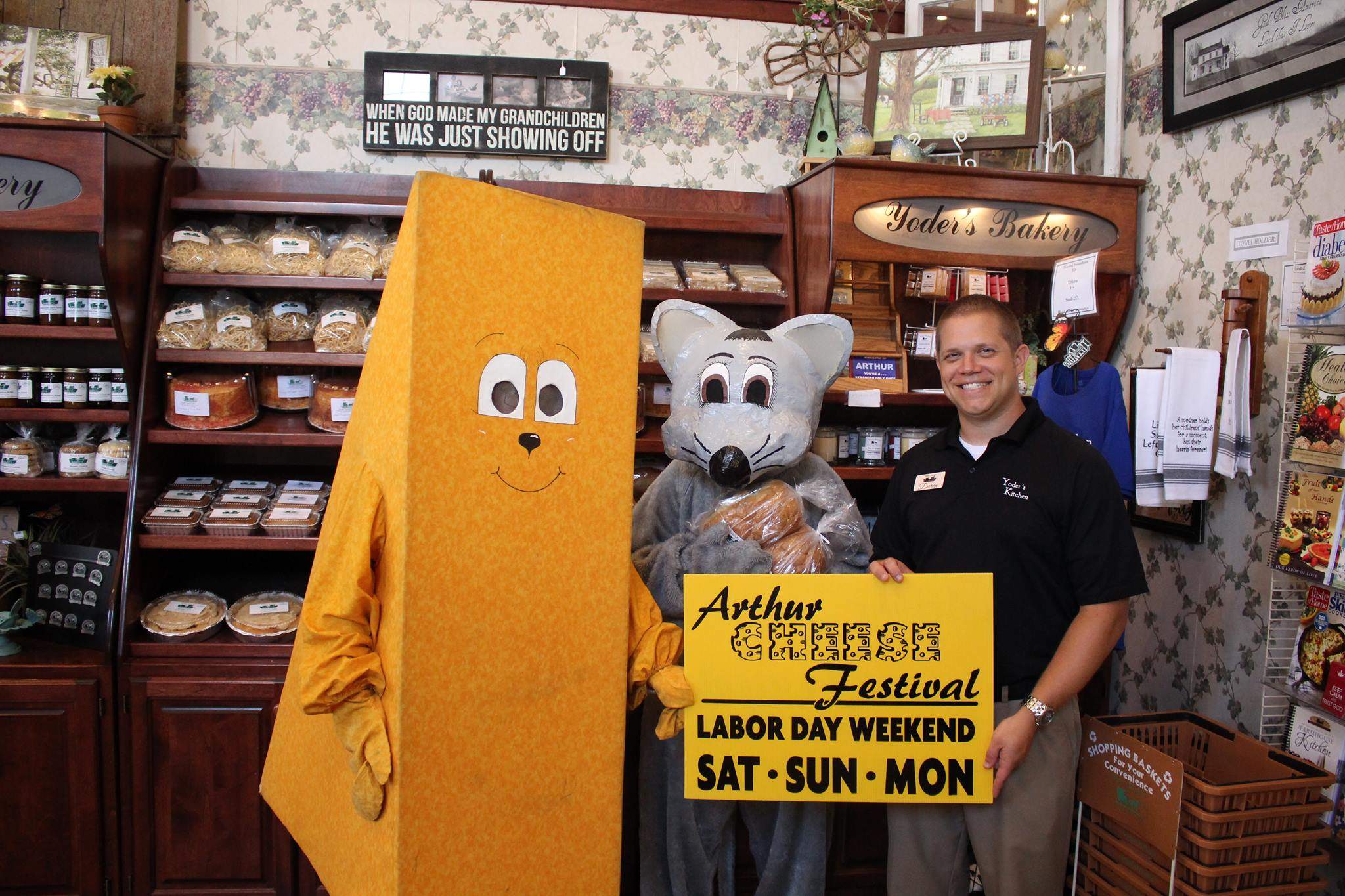 Mascottes en costume de fromage et en costume de souris à côté d'un homme tenant un panneau du festival Arthur Cheese.
