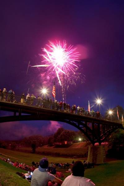 Personnes assises dans un parc le long d'un pont, regardant les feux d'artifice dans le ciel
