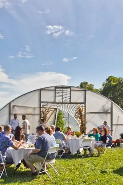 Des groupes de personnes assises autour de tables en plein air, au soleil, dégustent de la nourriture et des boissons.