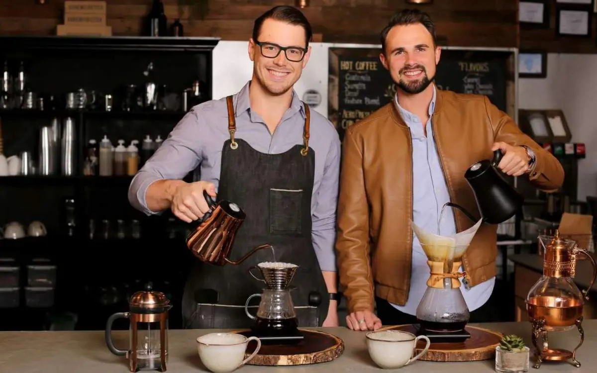 Deux hommes regardent la caméra tout en versant du café filtre dans un café.