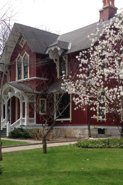 Une grande maison en bois rouge du XIXe siècle, à deux étages, derrière une pelouse et un arbre à fleurs.