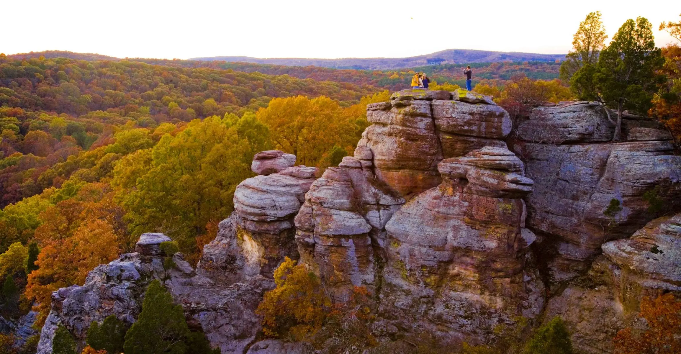 Un groupe éloigné de quatre personnes posant pour une photo au sommet de falaises rocheuses, surplombant des forêts d'arbres verts et dorés.