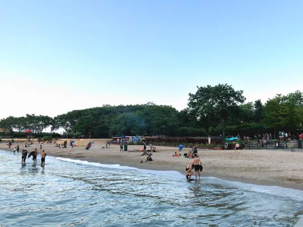 Petits groupes de personnes sur une plage tranquille au bord d'un lac au crépuscule, avec des arbres verts derrière eux.