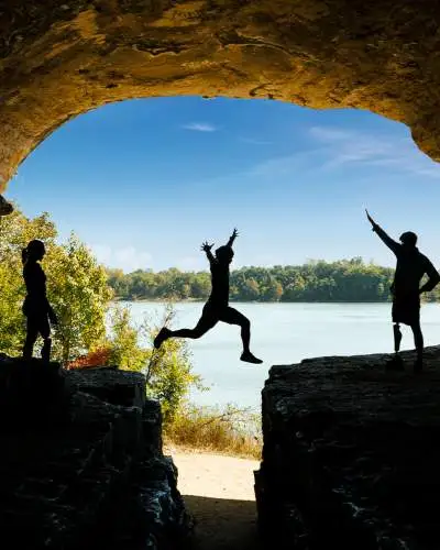 Une personne saute sur un rocher avec des amis, le ciel bleu et l'eau en arrière-plan.