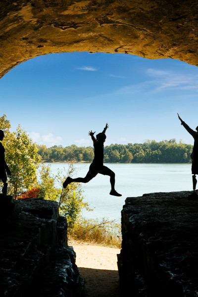 Une personne saute sur un rocher avec des amis, le ciel bleu et l'eau en arrière-plan.