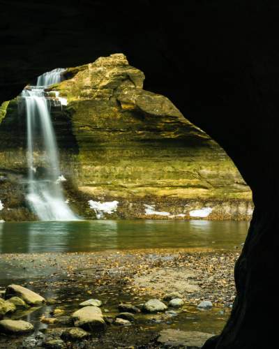 En regardant vers l'extérieur d'une grotte, une cascade se jette dans un bassin, à côté de parois rocheuses tapissées de neige.