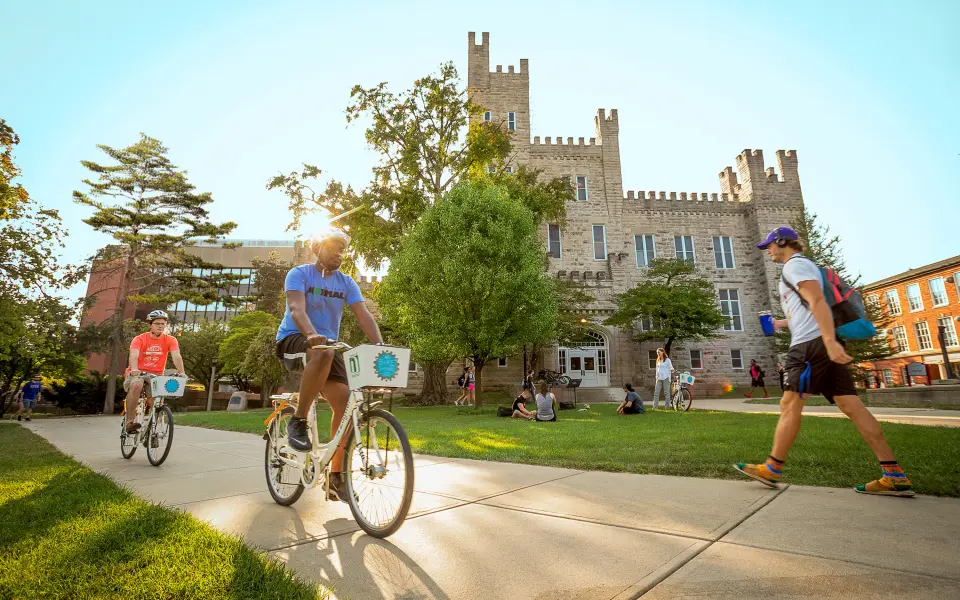 Deux personnes traversent à vélo un campus universitaire par une journée ensoleillée.