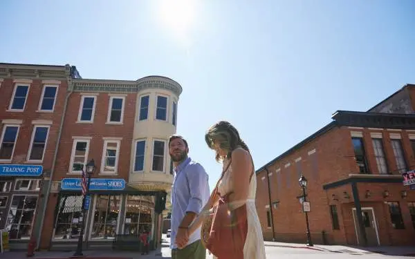 Plan en contre-plongée d'un jeune couple marchant dans la rue principale d'une petite ville, avec des bâtiments en briques et le soleil derrière eux.