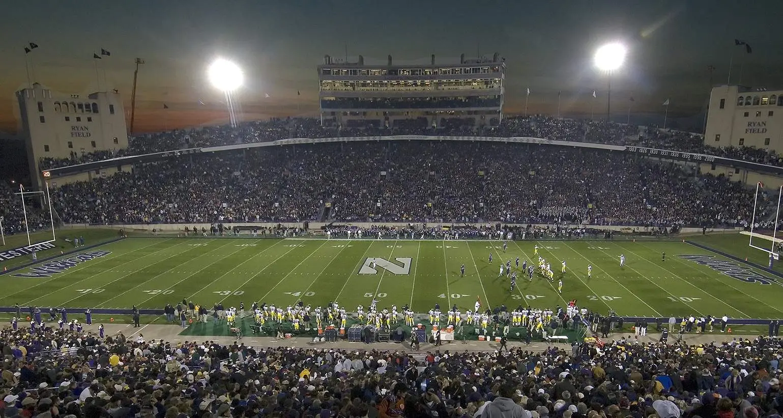 Un grand stade de football universitaire, la nuit précédant un match, avec des tribunes pleines de spectateurs.