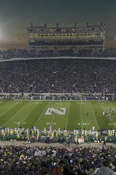 Un grand stade de football universitaire, la nuit précédant un match, avec des tribunes pleines de spectateurs.