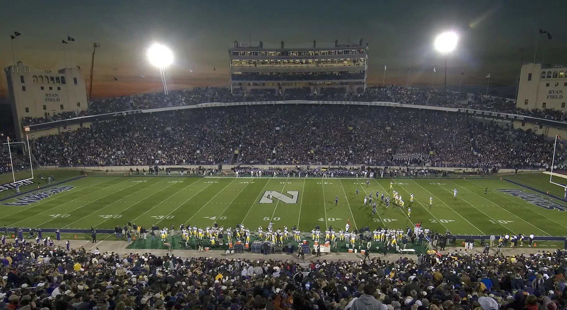 Un grand stade de football universitaire, la nuit précédant un match, avec des tribunes pleines de spectateurs.