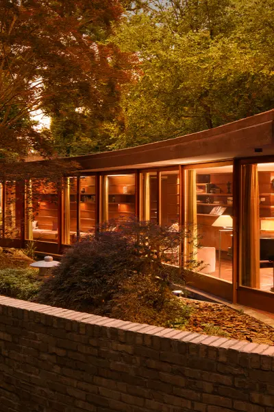 Une maison moderne aux vitres incurvées, illuminée à la tombée de la nuit, entourée d'arbres et d'un muret en briques.