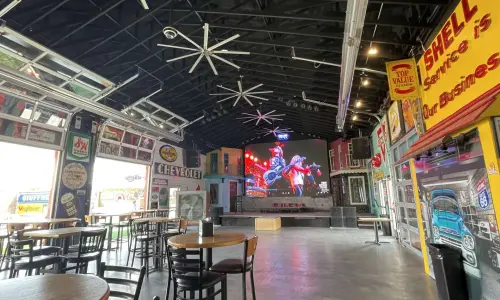 Intérieur d'un bar spacieux proposant des concerts, avec des tables, des enseignes et des éléments de décoration inspirés de l'automobile vintage, ainsi qu'un grand écran au-dessus de la scène.